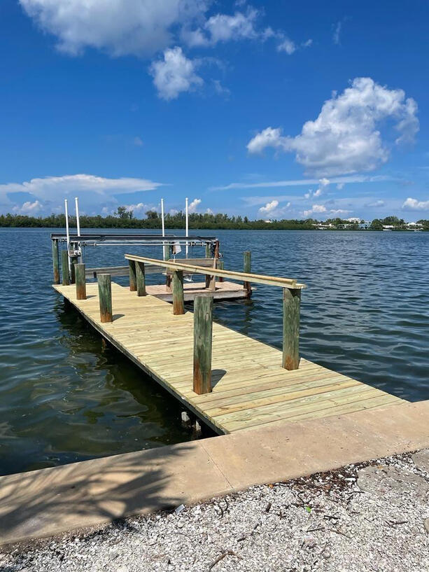 A beautifully constructed wooden dock extends over calm, blue-green waters under a vibrant blue sky dotted with fluffy white clouds. The dock features sturdy pressure-treated wood planks with reinforced posts and railings, providing safe and secure access