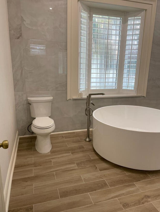 Spa-Inspired Bathroom Retreat This elegant bathroom features a freestanding soaking tub with modern floor-mounted faucet, warm wood-look tile flooring, and a soft gray marble-style wall finish. The custom bay window with plantation shutters allows natural
