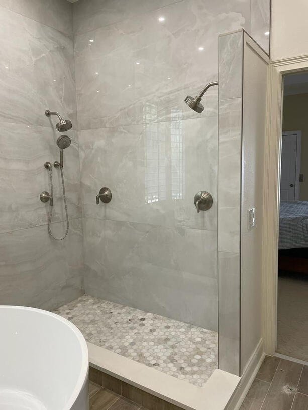 A luxurious, contemporary walk-in shower with a dual showerhead setup, featuring sleek brushed nickel fixtures. The walls are clad in glossy, large-format marble-look porcelain tiles with soft grey veining, creating a clean and sophisticated aesthetic. The
